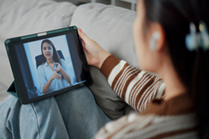 Selective focus, female patient sitting on sofa receiving online therapy from female medical professional on digital tablet in living room at home. Mental health care, Remote counselling
